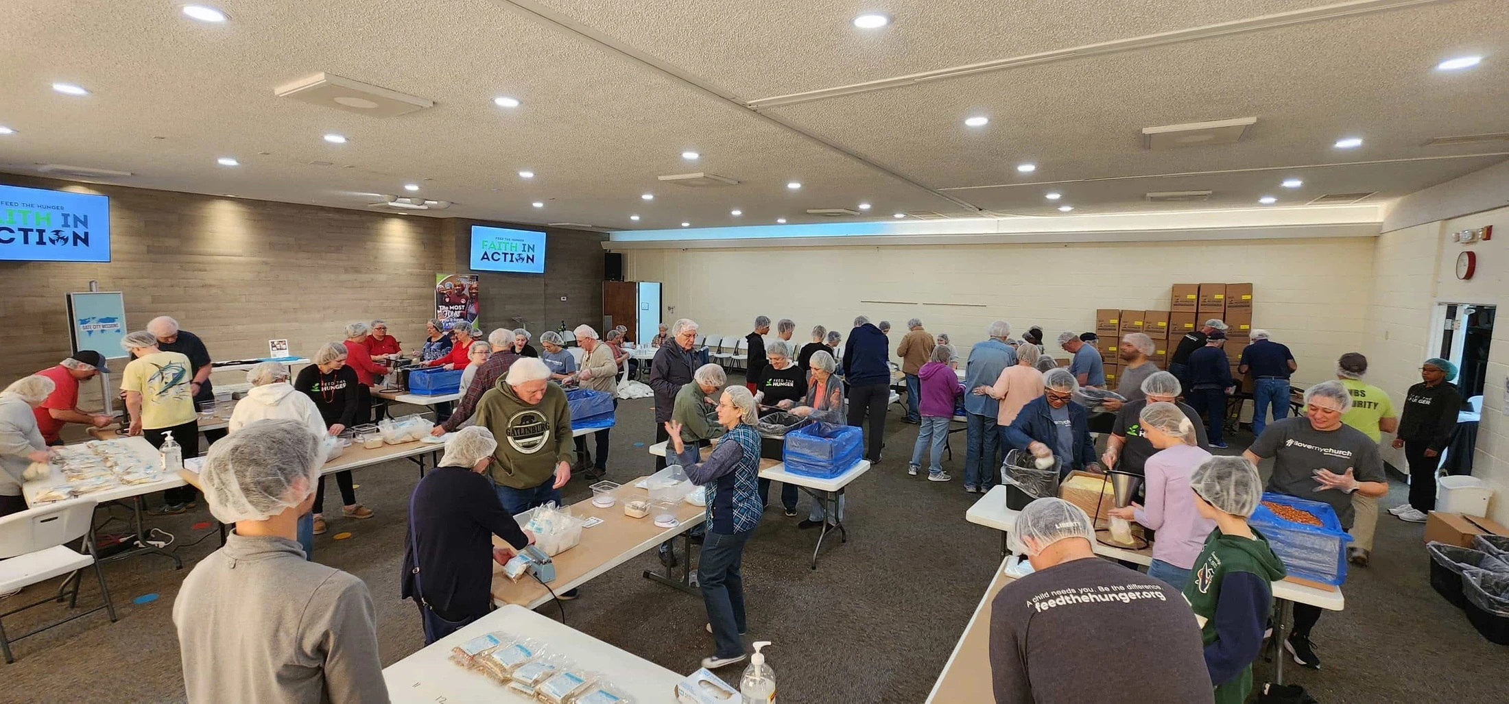 A large group of volunteers wearing hairnets work together at long tables, packing meals in a spacious hall for a hunger relief charity event.