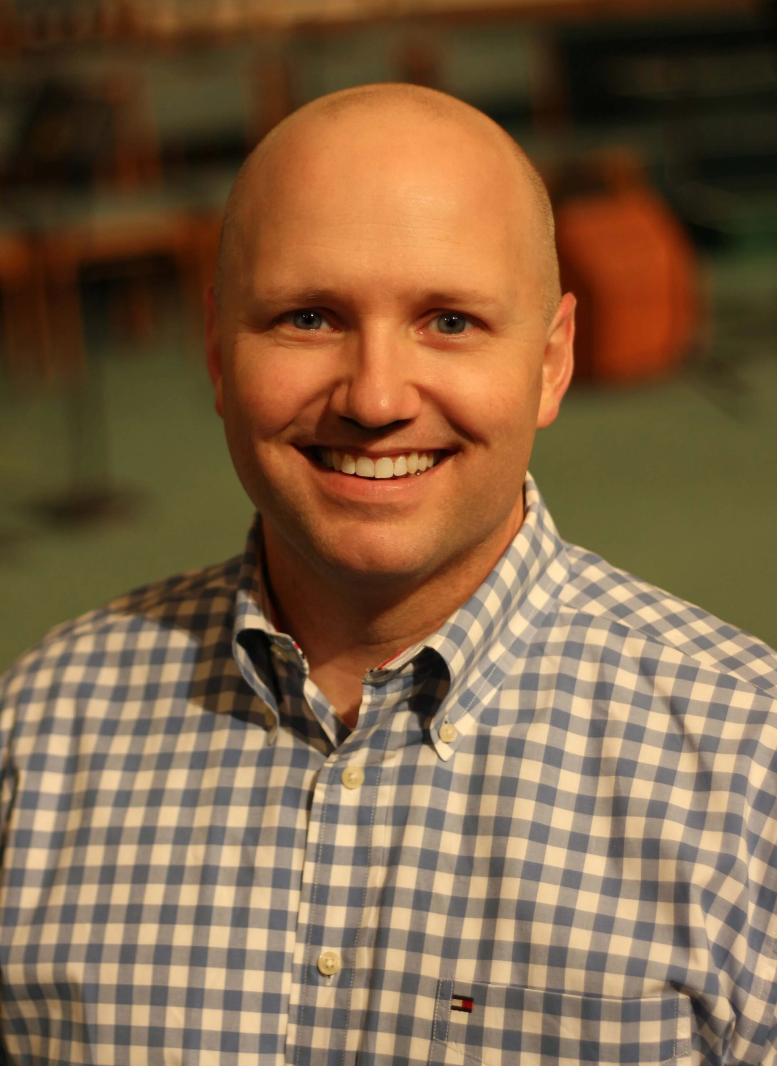 A head-and-shoulders portrait of a bald, blue-eyed man smiling warmly at the camera. He is wearing a blue and white checkered button-down shirt.