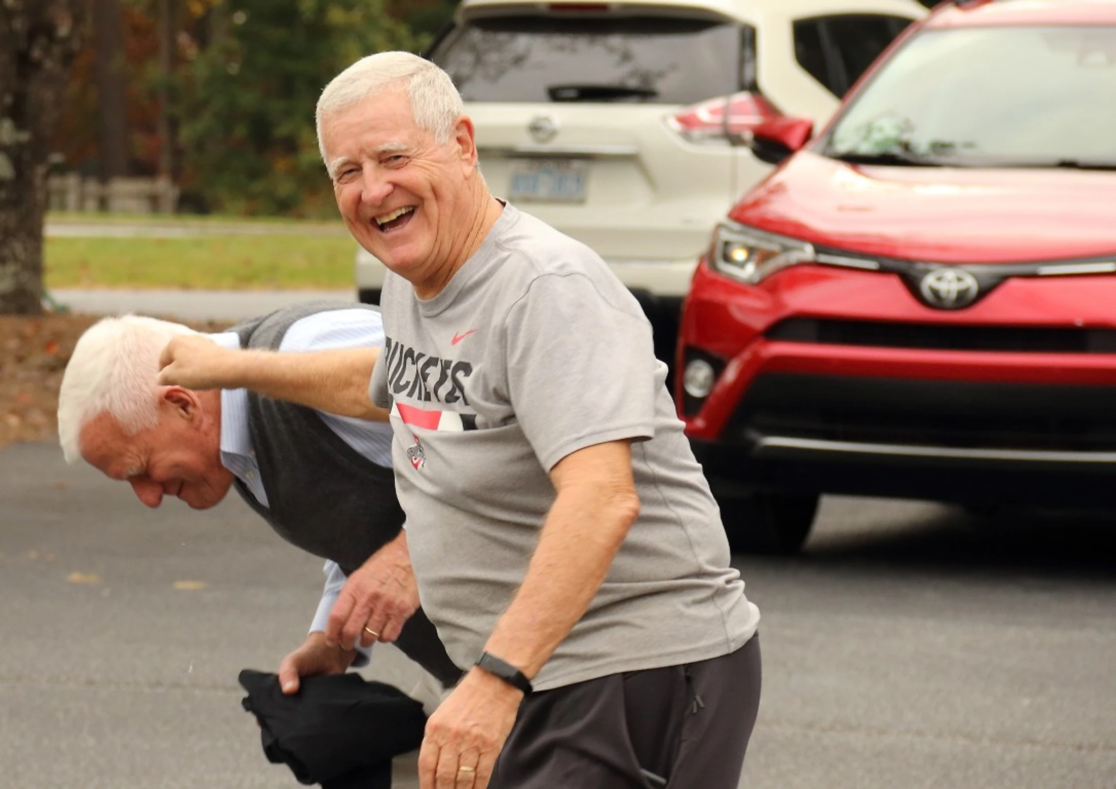 An older man in a grey shirt laughs as he playfully punches another man on the back of the head. They are outdoors in a parking lot with cars behind them.