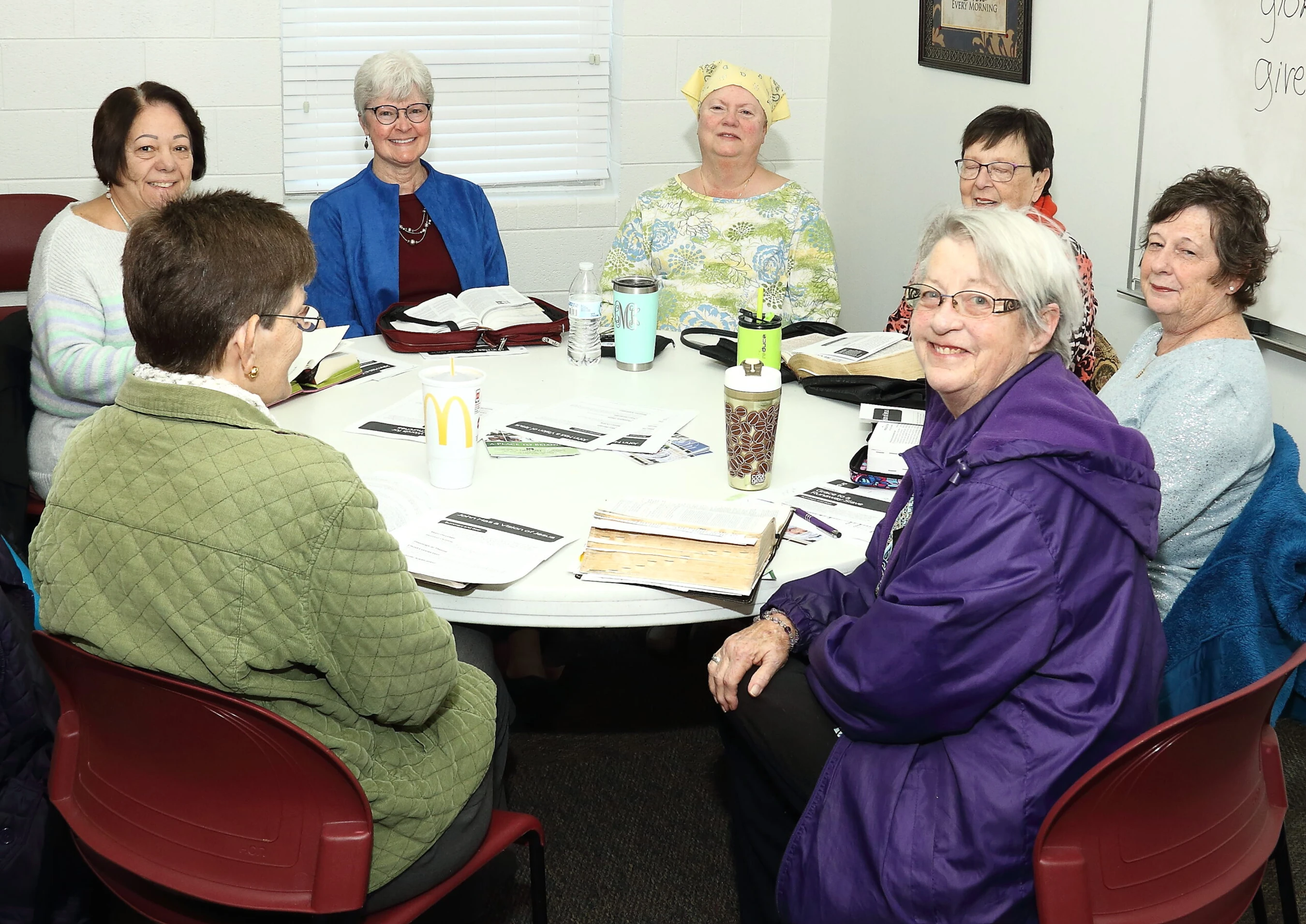 A group of seven older women smile for the camera while seated around a white table in a classroom for a study group meeting.