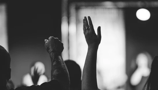 A black and white photo showing the backs of people raising their hands. In the foreground, a man's tattooed arm and a woman's hand are lifted.