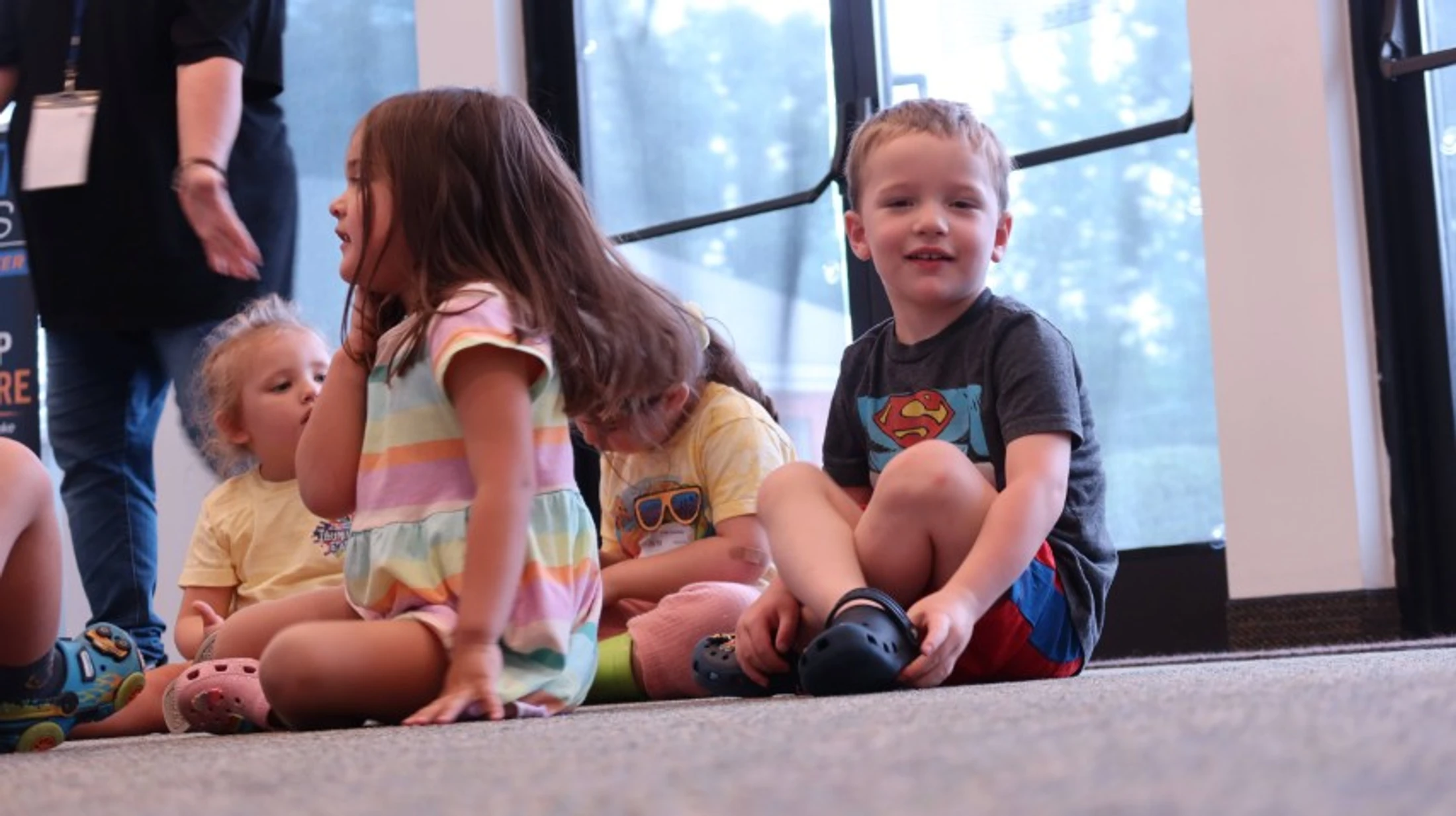 A low-angle shot of several young children sitting on a carpeted floor. A boy in a Superman shirt smiles at the camera while others sit beside him.