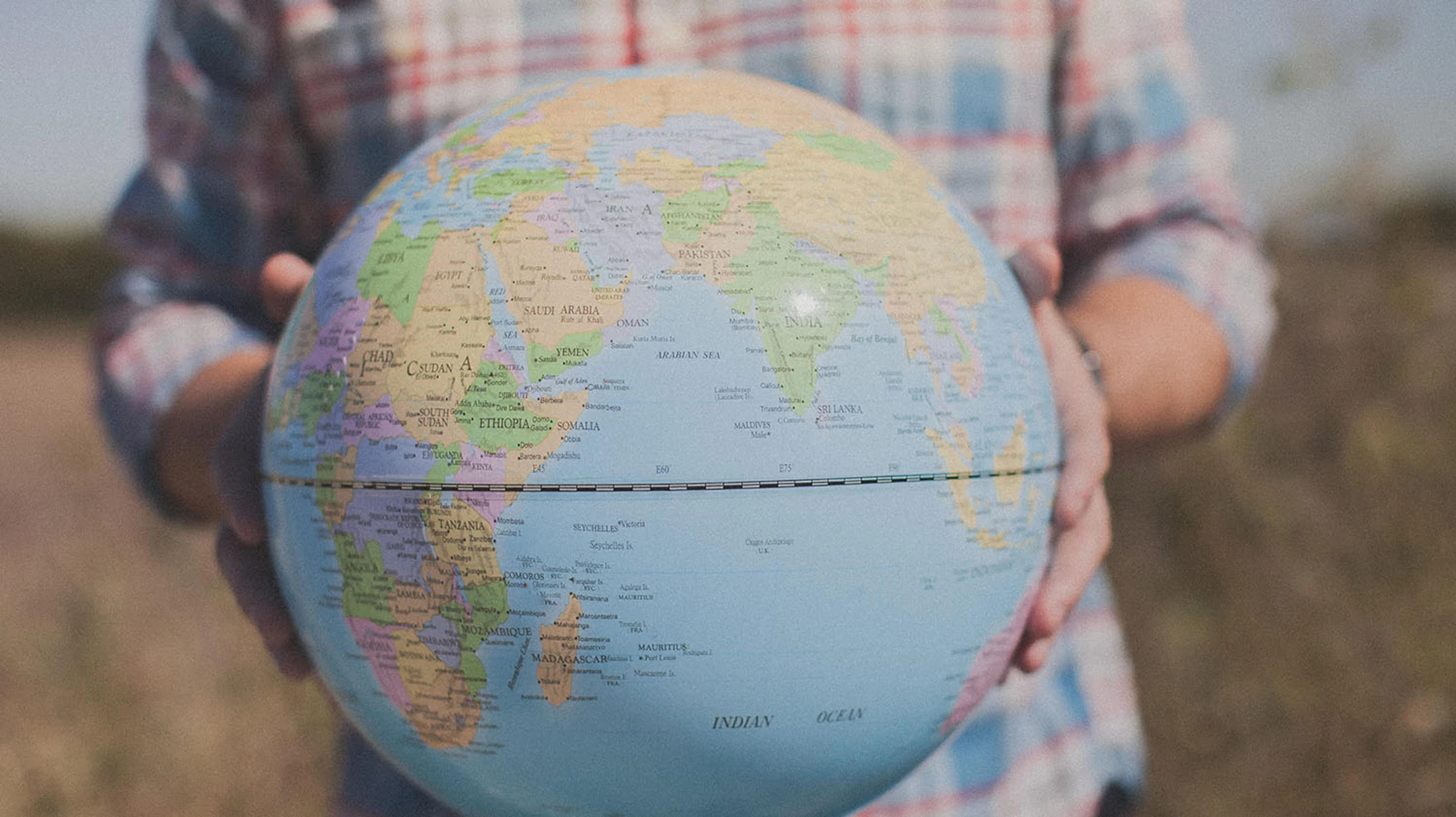 A person in a plaid shirt holds up a world globe, with Africa, the Middle East, and the Indian Ocean in focus. The background is softly blurred.