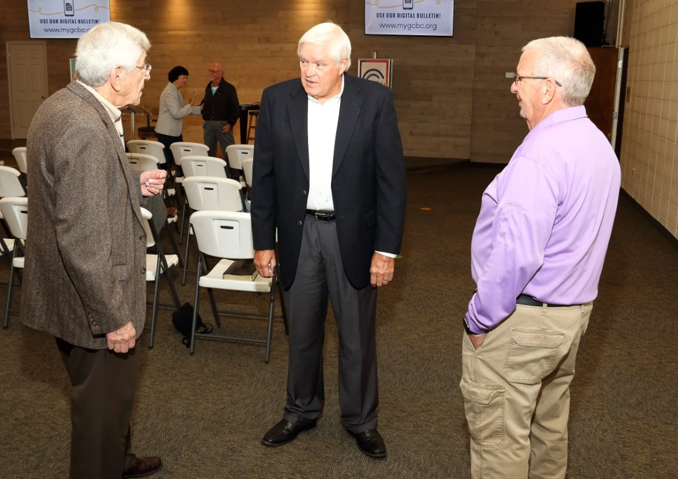 Three older men stand talking in a room with rows of white folding chairs. Two wear jackets and one wears a purple shirt. Other people are in the background.