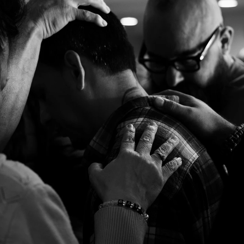 A black and white photo showing several people gathering around a man, placing their hands on his head and shoulder in a gesture of prayer or support.