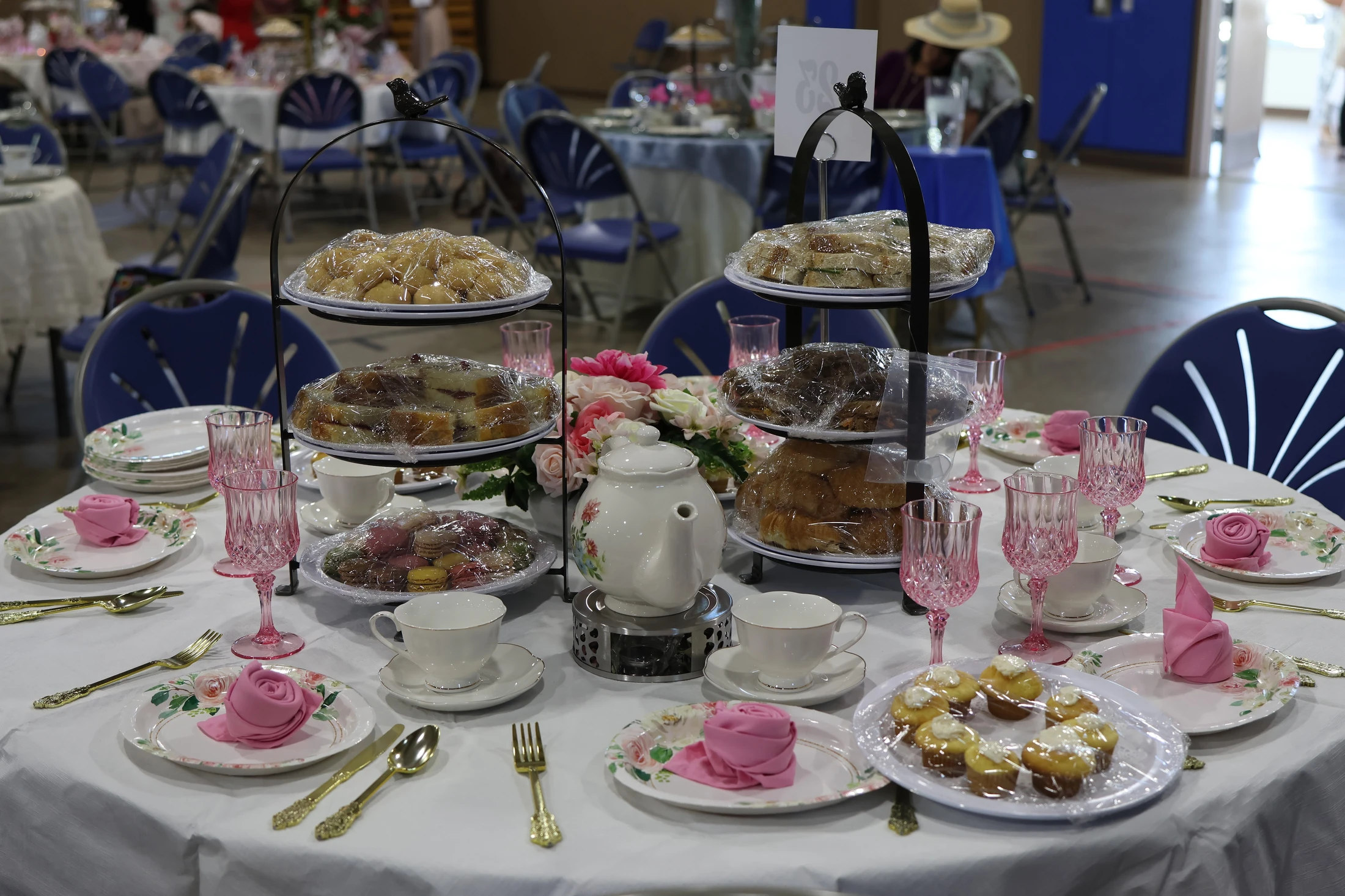 A round table set for a tea party with floral plates, pink glasses, and tiered stands full of plastic-wrapped sandwiches, cakes, and pastries.