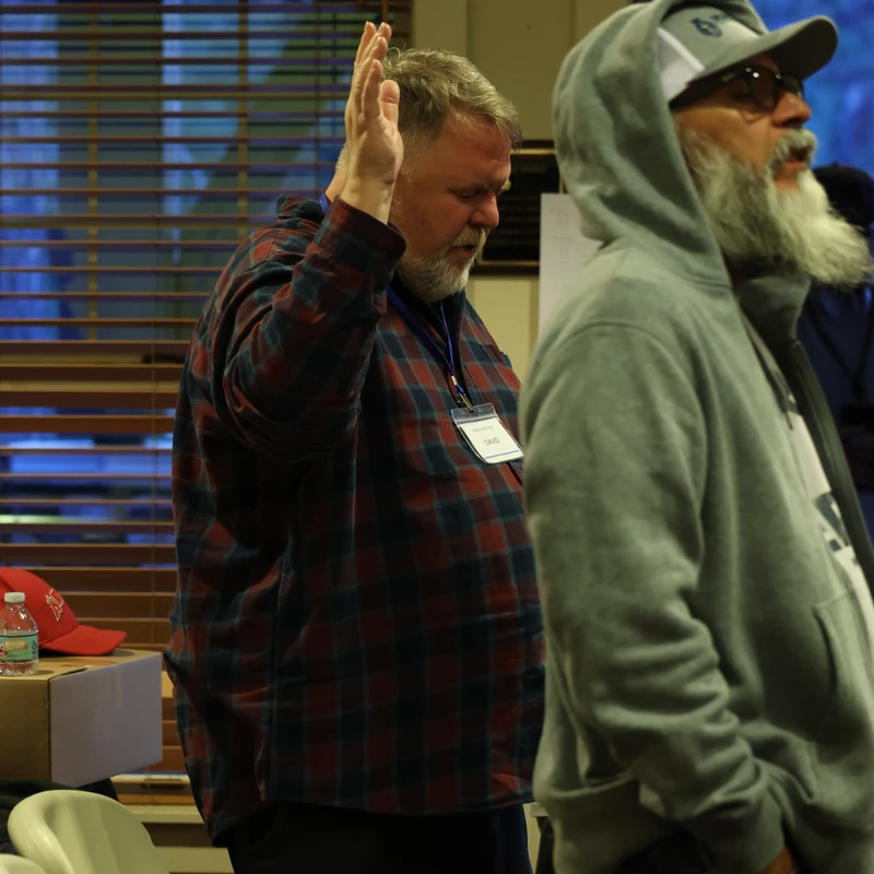 A man in a plaid shirt stands with his eyes closed and hand raised in worship. Next to him, another man with a long white beard and a hoodie looks on.
