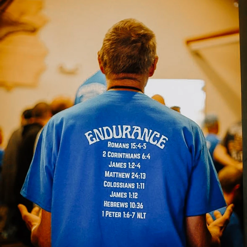A man viewed from behind wearing a blue t-shirt with the word 'ENDURANCE' and a list of related Bible verses printed in white on the back.
