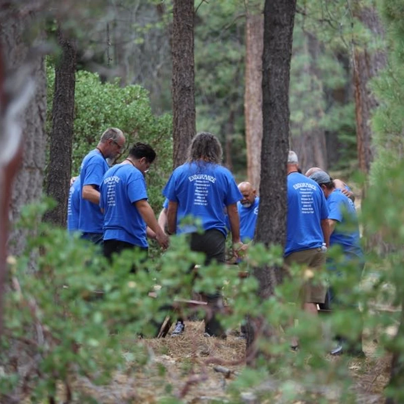 A group of men in matching blue t-shirts stand in a circle holding hands with their heads bowed in prayer, seen through trees in a forest.