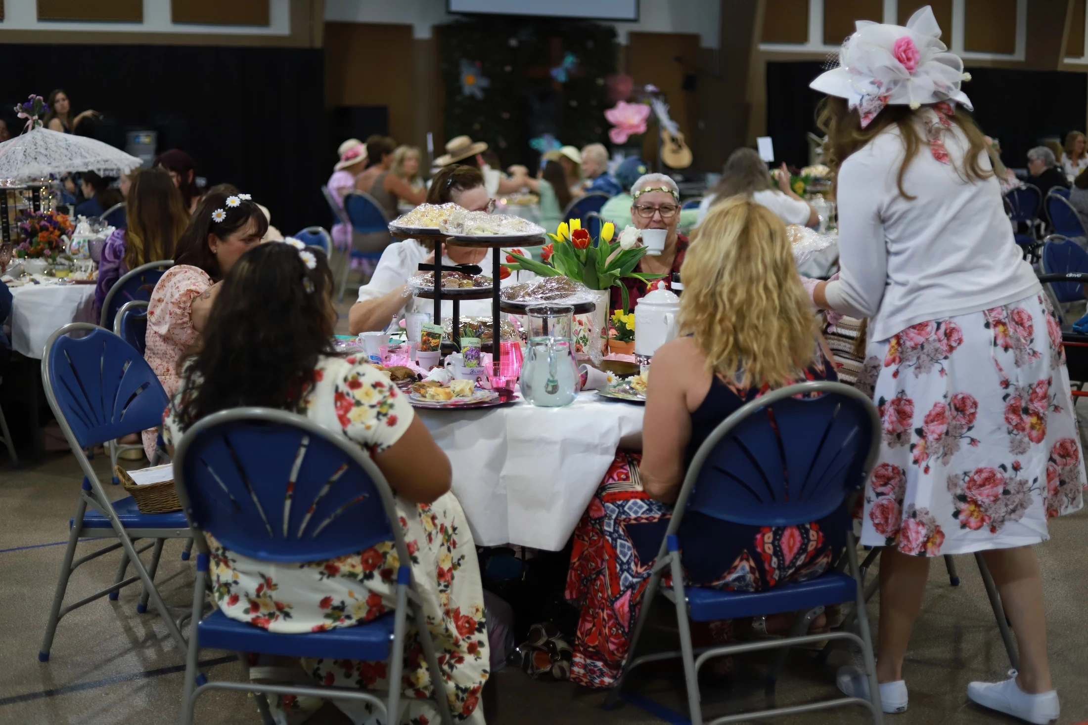 A group of women in floral dresses and hats sit around a decorated table enjoying food and conversation at an indoor tea party event.