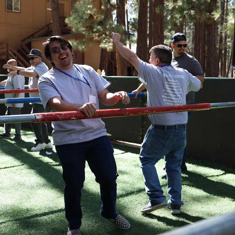 A group of men laugh and cheer while playing human foosball outdoors at a sunny, wooded camp, holding onto bars on an artificial turf field.