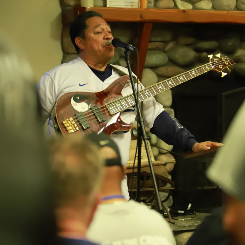 A man with a mustache plays a bass guitar and sings into a microphone during an indoor performance in front of a stone fireplace.