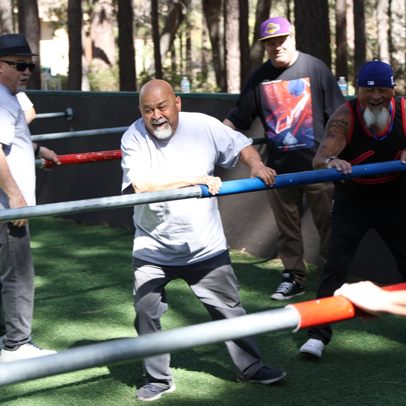 A group of men laugh and smile while playing a competitive game of life-sized human foosball on an outdoor turf field surrounded by trees.