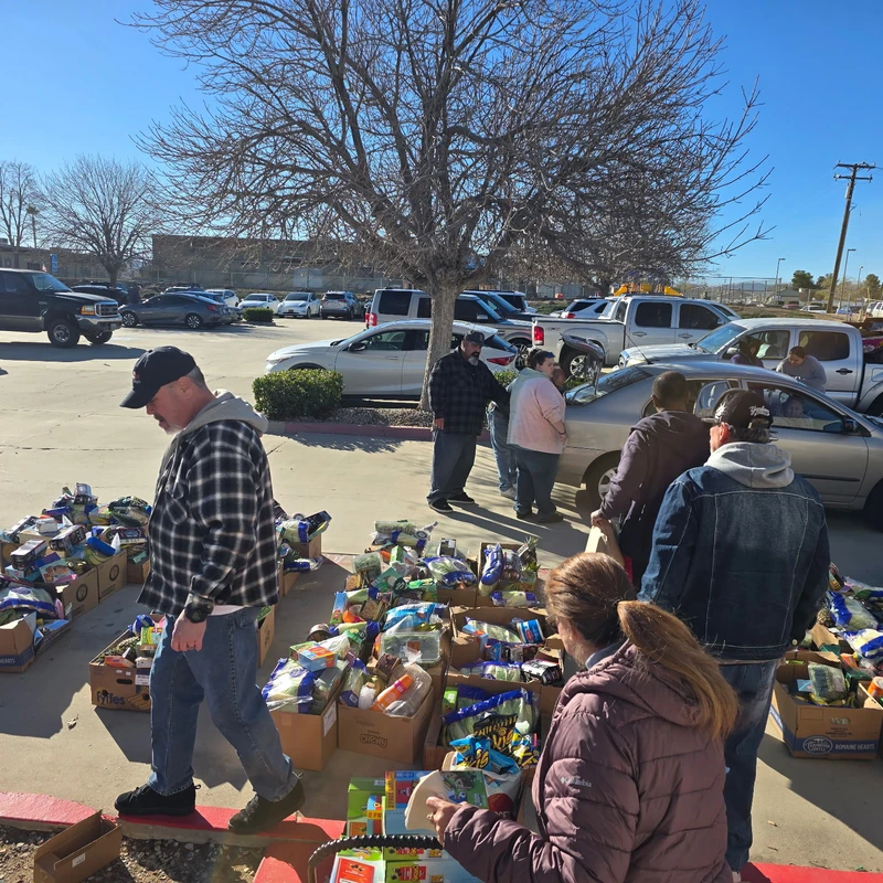 A group of people gathers in a sunny parking lot to select groceries from cardboard boxes lined up on the curb during a food drive.