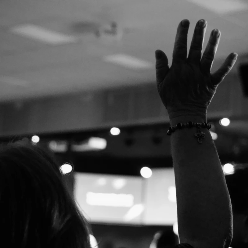 A black and white photo from behind a person with their hand raised. They wear a bracelet with a cross charm, and the background is a blurry indoor setting.