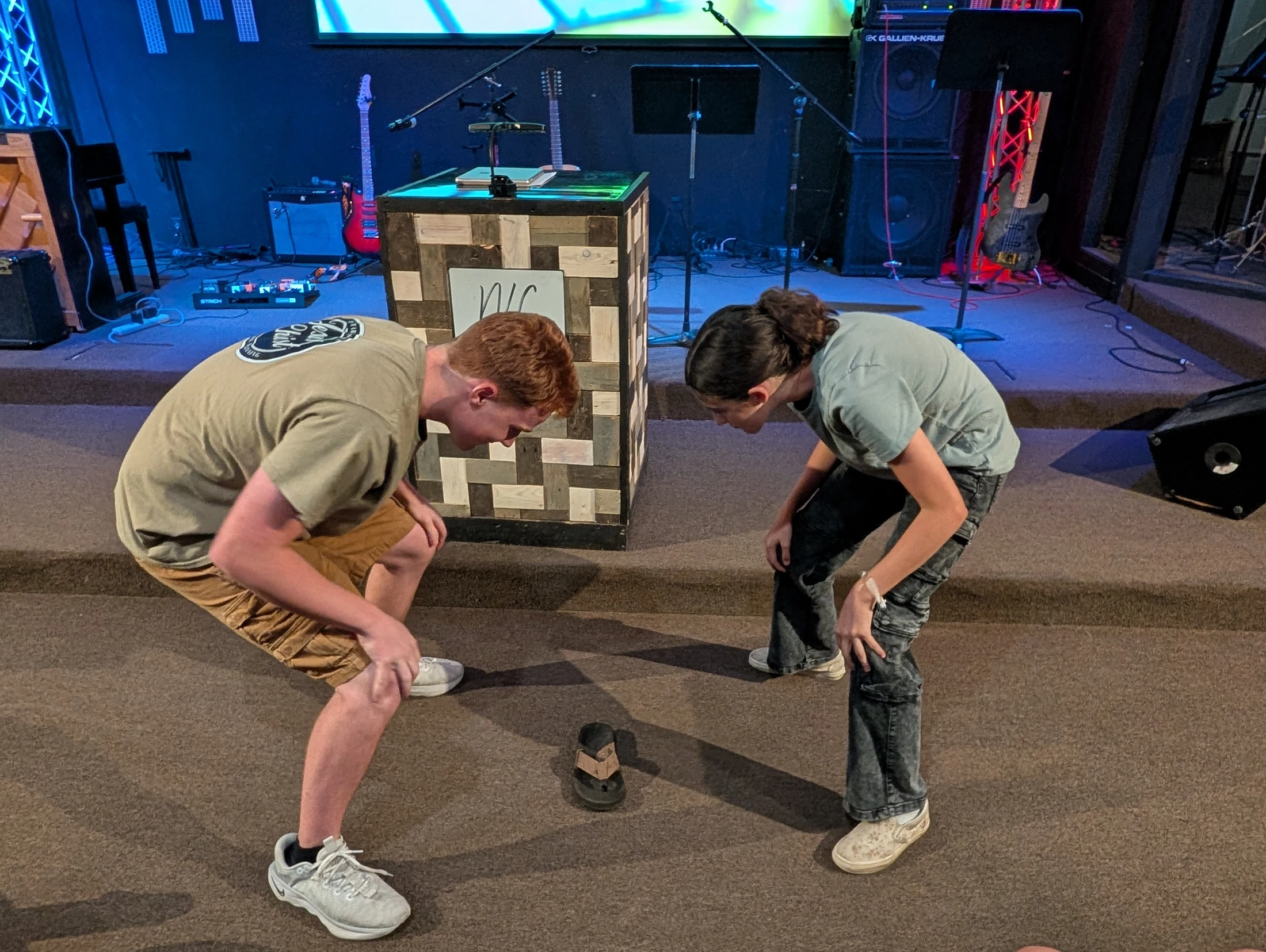 Two young men bend over, looking intently at a single black flip-flop on the carpeted floor of a stage with musical equipment in the background.