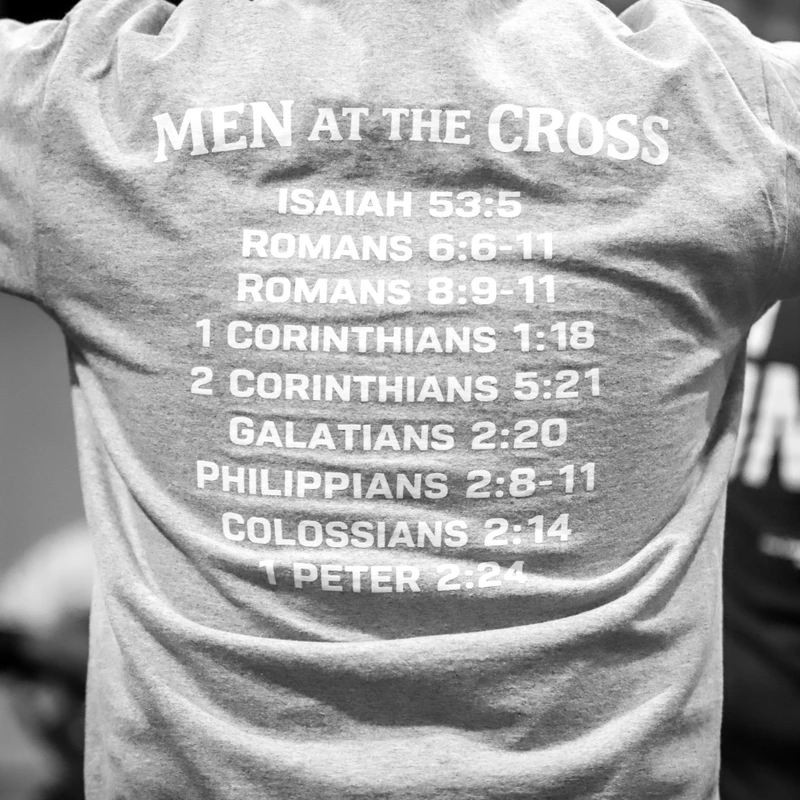 Black and white photo of a man's back wearing a grey t-shirt with 'MEN AT THE CROSS' and a list of Bible verses printed on it.