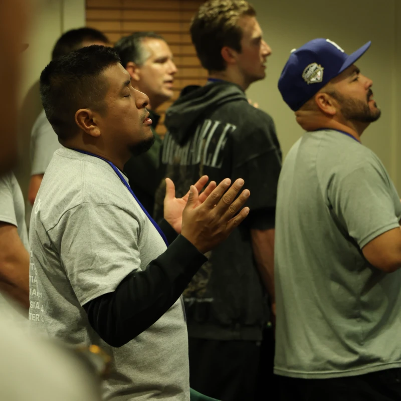 A man in a grey shirt stands with his eyes closed and hands raised in prayer, surrounded by other men at an indoor conference or worship service.