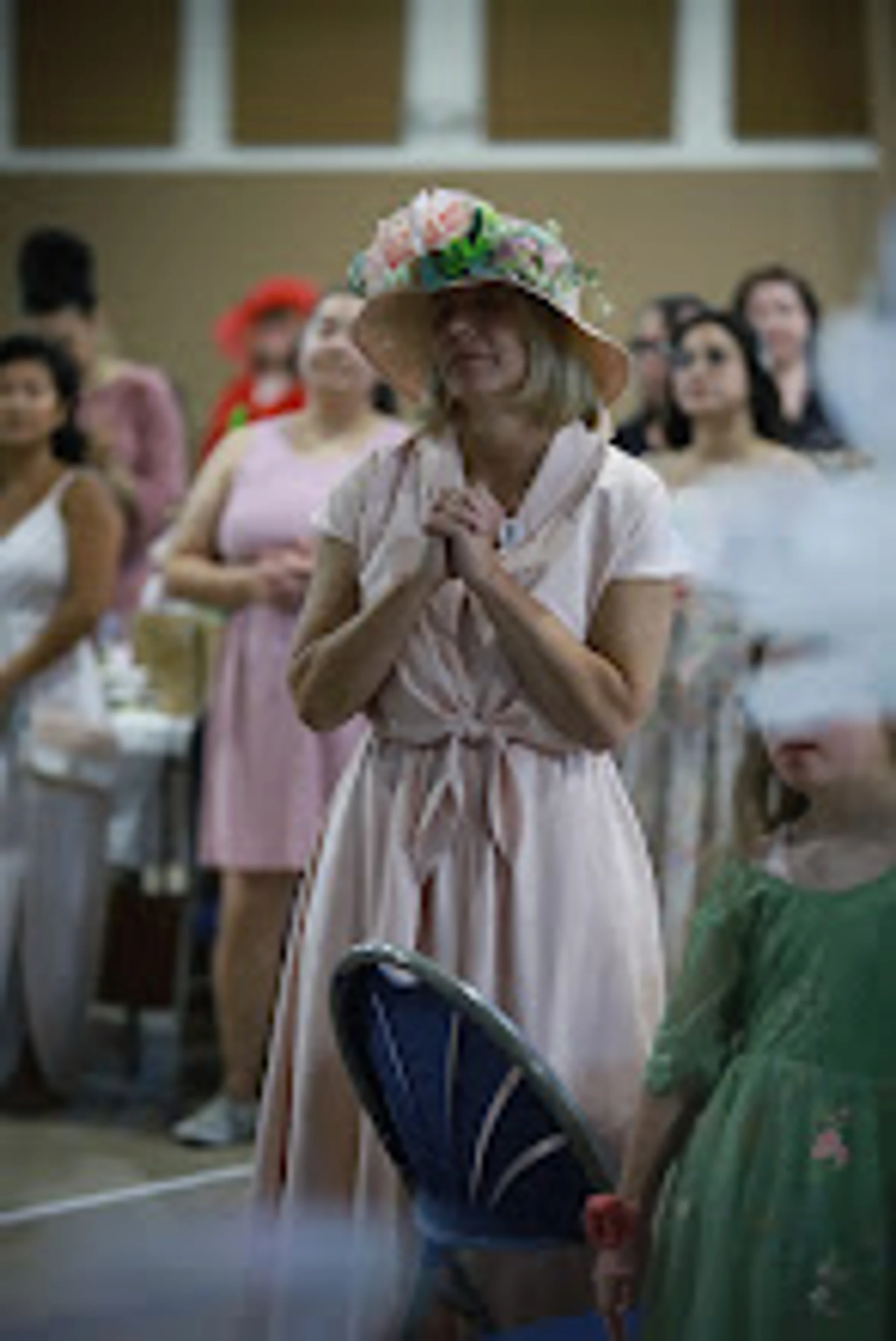A woman in a light pink dress and a straw hat adorned with pink flowers stands with her hands clasped, looking upward with a serene expression during an event.