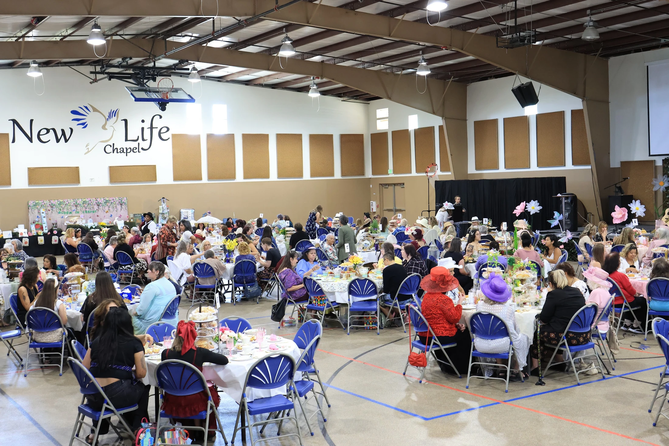 A wide shot of a large church gymnasium filled with women sitting at decorated round tables for a festive tea party luncheon, many wearing hats.