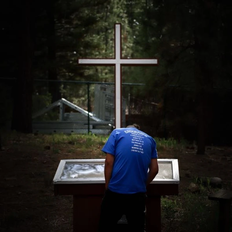 A person seen from behind in a blue shirt stands with their head bowed at a lectern in a dark forest. A large white Christian cross is behind them.