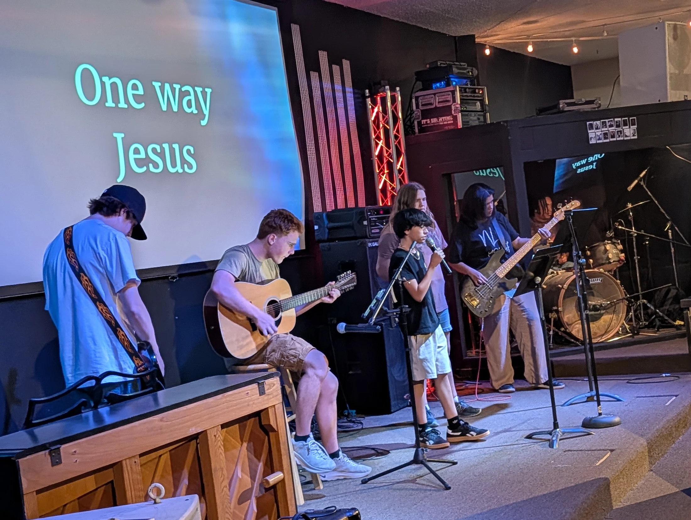A group of young musicians perform on a dimly lit stage. They include a singer, guitarist, and bassist, with a screen behind them reading 'One way Jesus'.