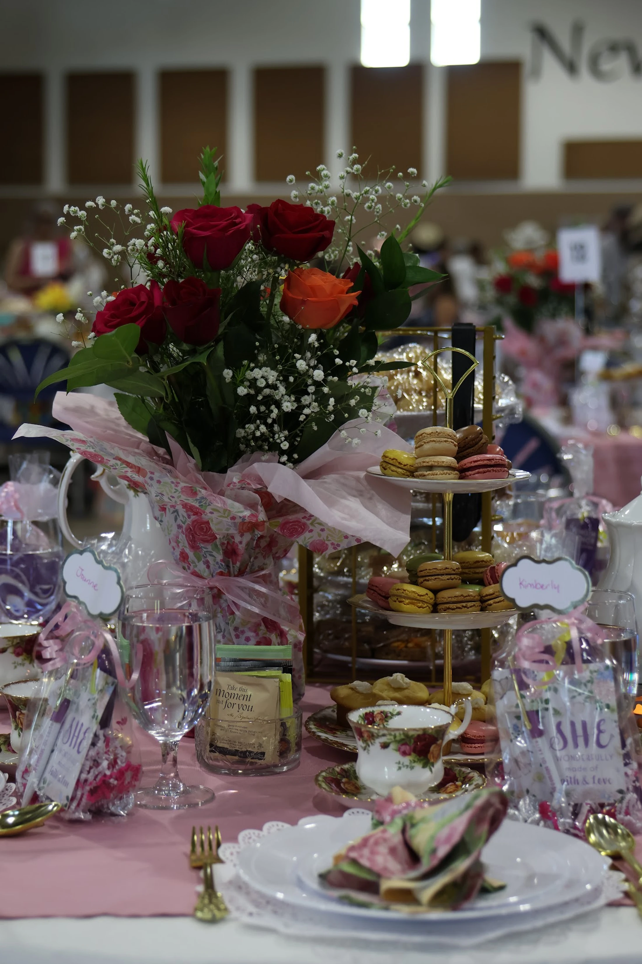 An elegant table setting for a tea party with a vase of red roses, a tiered stand of macarons, and floral china on a pink tablecloth.