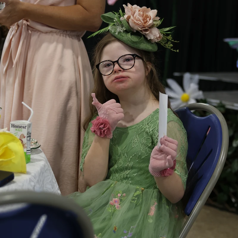 A young girl with Down syndrome in a green dress, floral hat, and pink gloves sits at a table. She looks at the camera with a pout and gives a thumbs up.