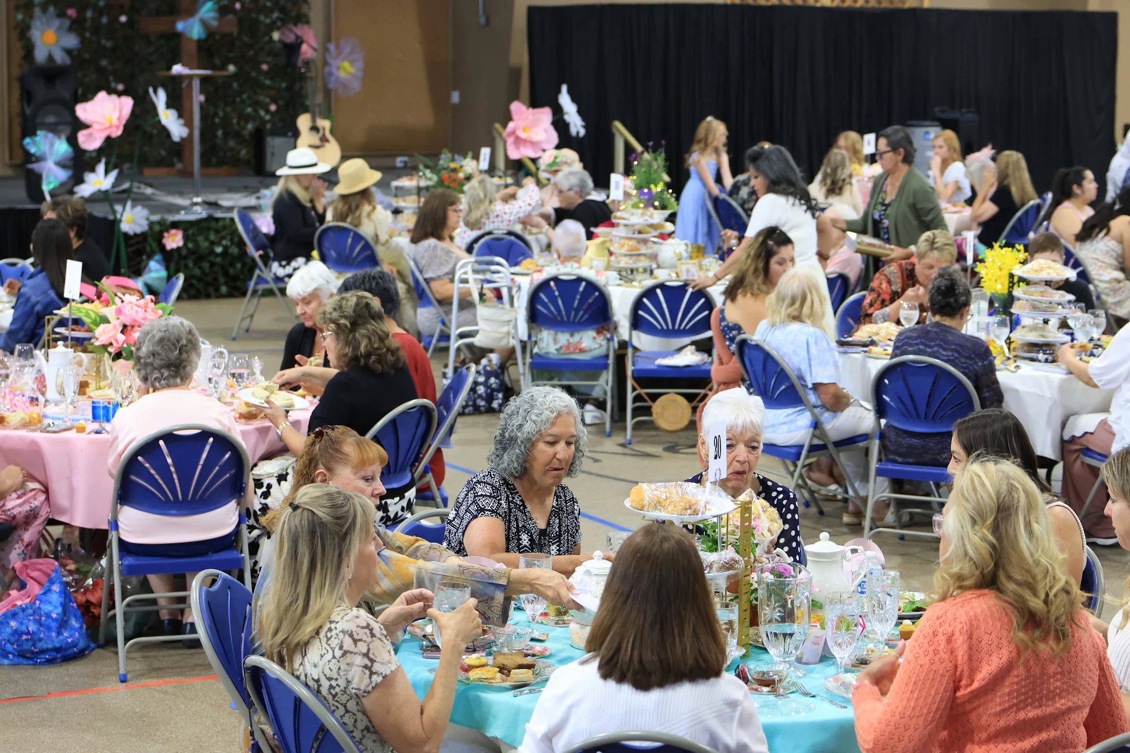 A large room filled with women of various ages seated at decorated round tables, enjoying food, drink, and conversation during a festive tea party event.