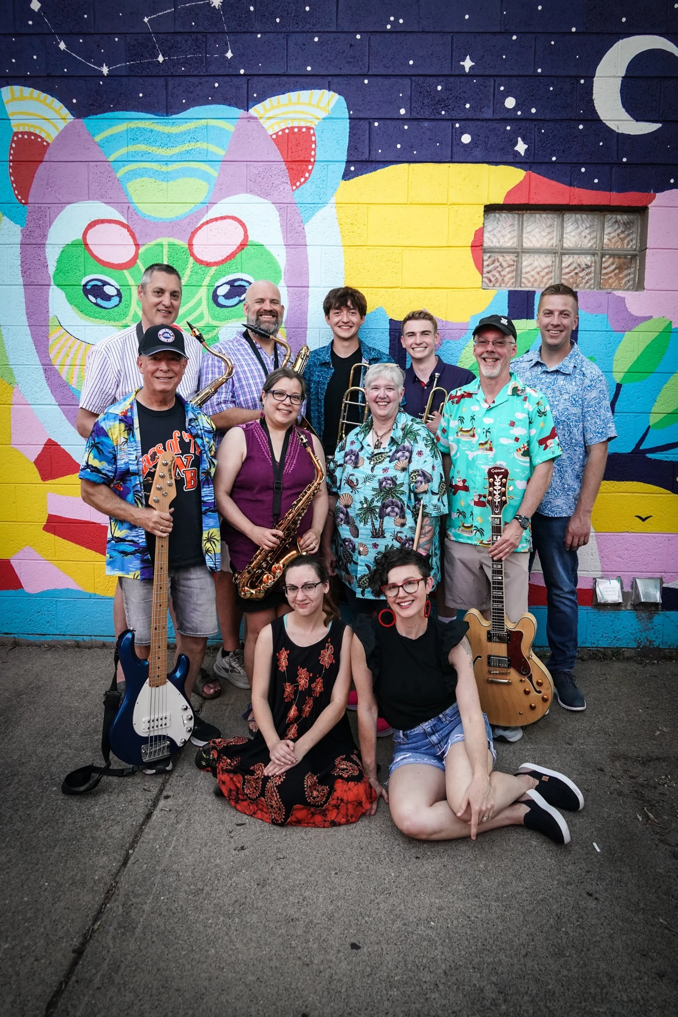 A band of eleven musicians holding instruments like guitars and saxophones poses for a group photo in front of a colorful, whimsical mural on a brick wall.