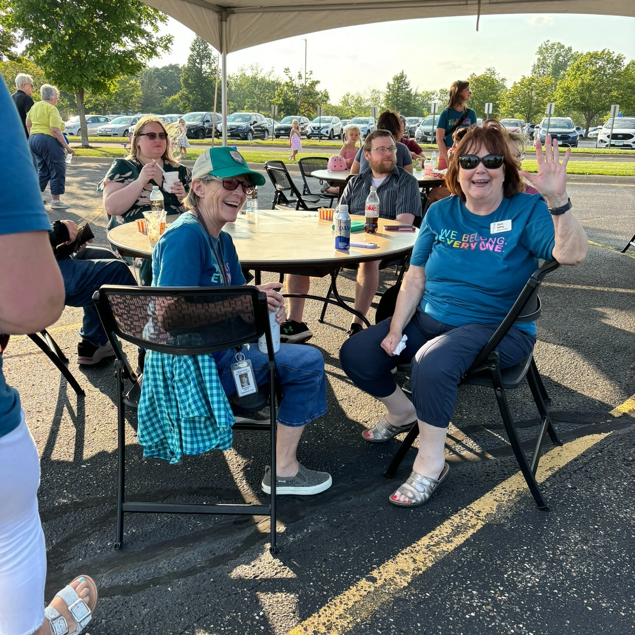 A group of people sit around a table under a tent in a sunny parking lot. One woman waves at the camera while another laughs.
