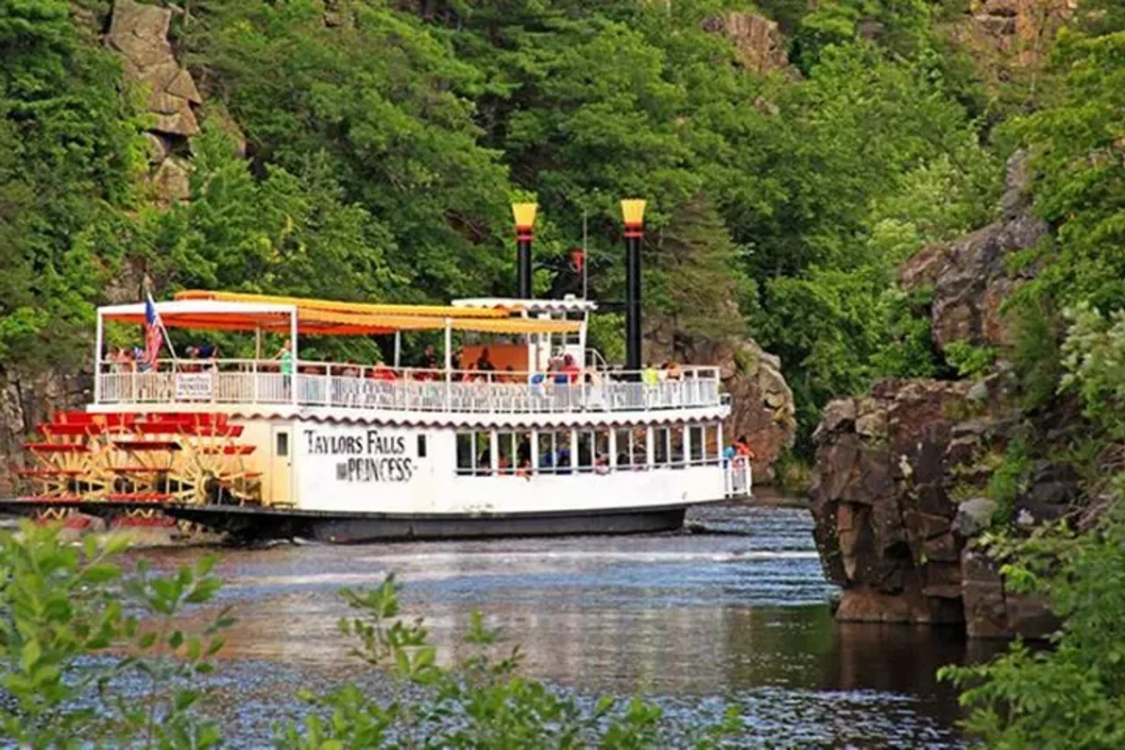 The Taylors Falls Princess, a white paddlewheel riverboat, carries tourists down a river surrounded by lush green trees and rocky cliffs on a sunny day.