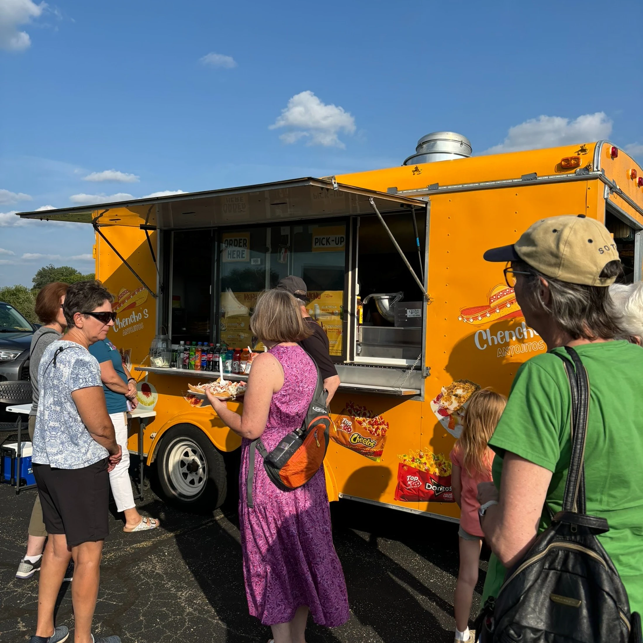 A group of people stand in line at a bright yellow food truck called Chencho's on a sunny day in a parking lot. A woman holds a plate of food.