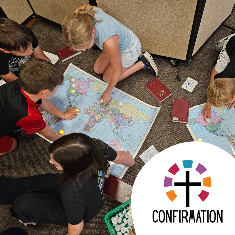 An overhead shot of several students in a confirmation class gathered on the floor, pointing to locations on a world map with Bibles and small lights nearby.