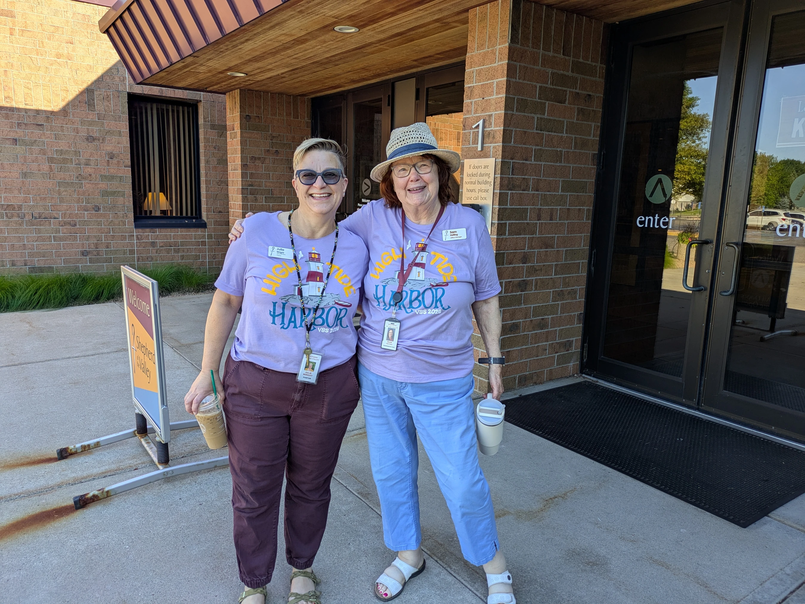 Two smiling women in matching purple 'High Tide Harbor VBS' t-shirts stand with arms around each other outside the entrance of a brick building.