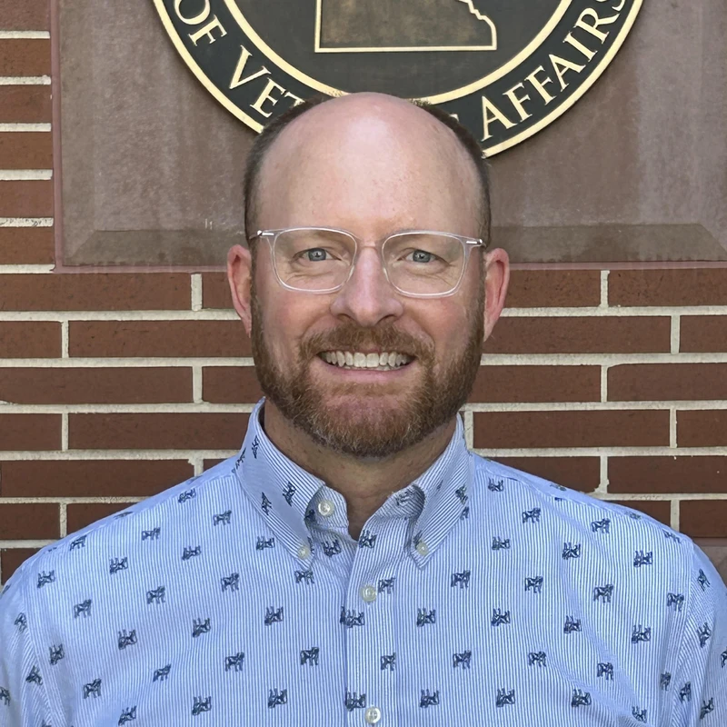 A smiling, bald man with a beard and glasses poses for a headshot. He is wearing a blue patterned shirt and standing in front of a brick wall with a seal.