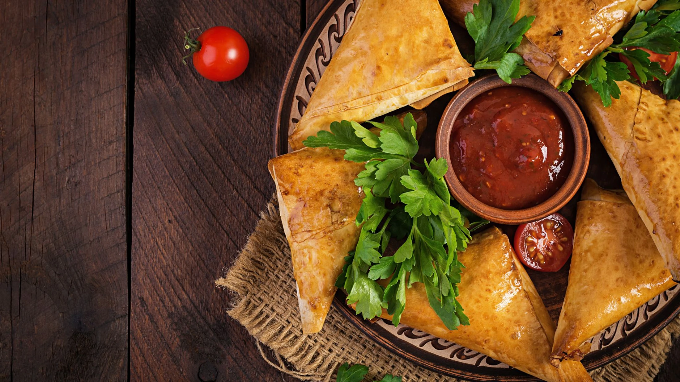 An overhead shot of a platter of golden-brown samosas with a small bowl of red dipping sauce, garnished with fresh parsley on a rustic wooden table.