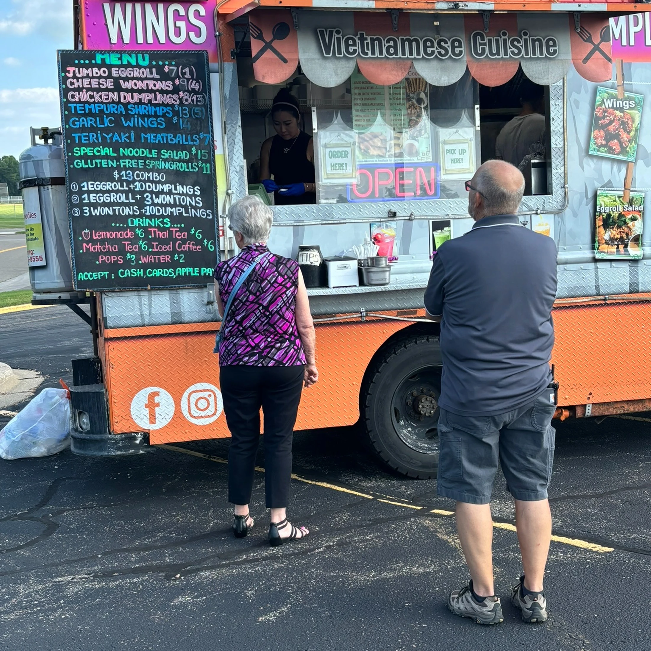An older man and woman stand in front of an orange food truck, reading the large chalkboard menu for Vietnamese cuisine.