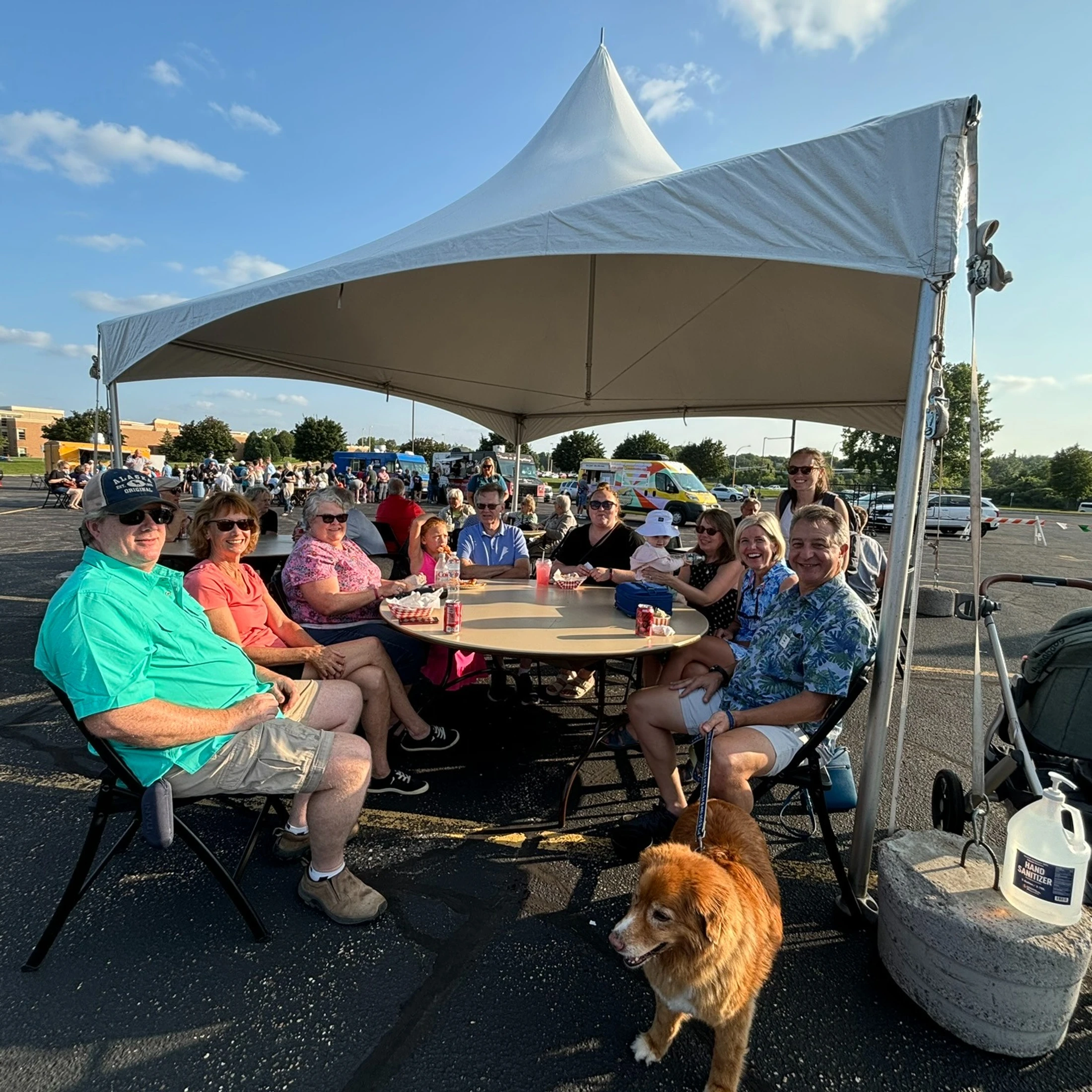 A group of people sitting around a table under a large white tent in a parking lot during a sunny day, with a brown dog in the foreground.