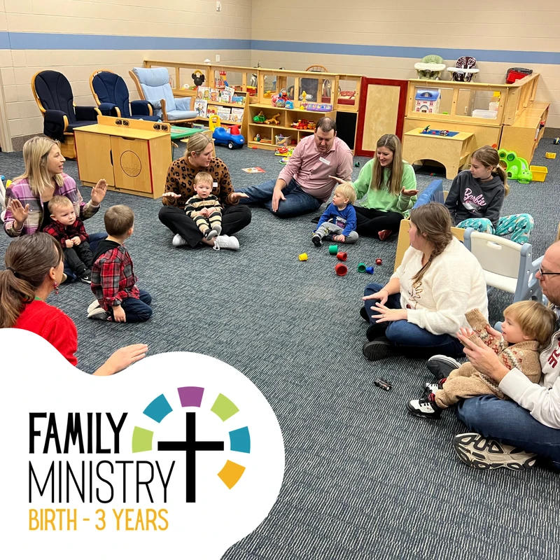 A group of parents and their young children sit in a circle on the floor of a playroom, participating in a family ministry class for ages birth to 3.