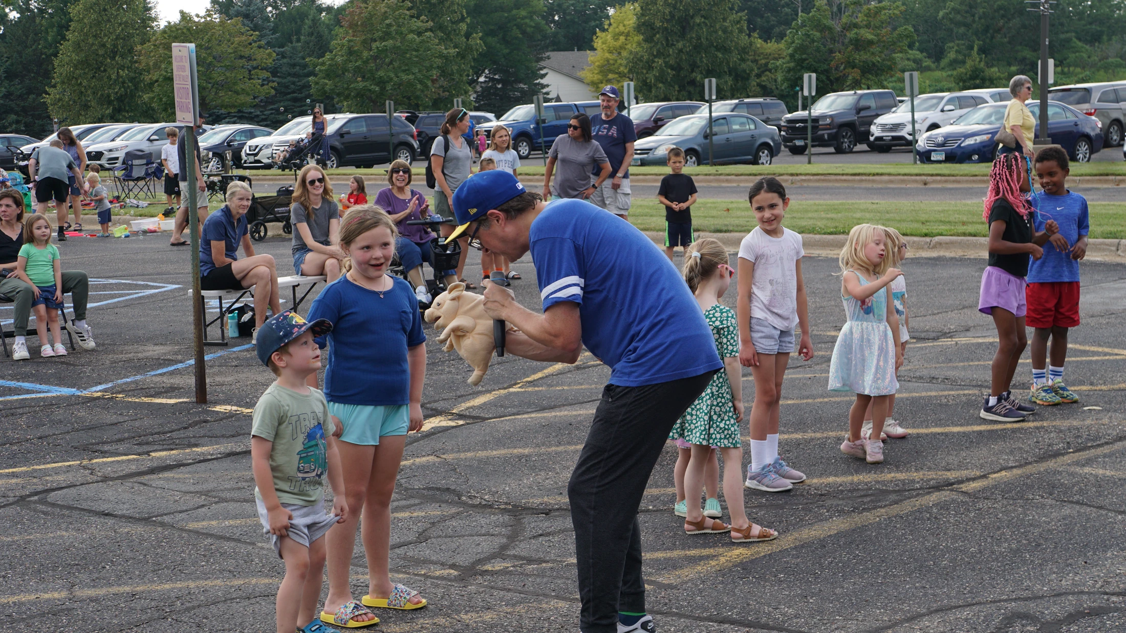 An entertainer in a blue shirt holds a pig puppet and a microphone, performing for a group of smiling children gathered in a parking lot for an event.