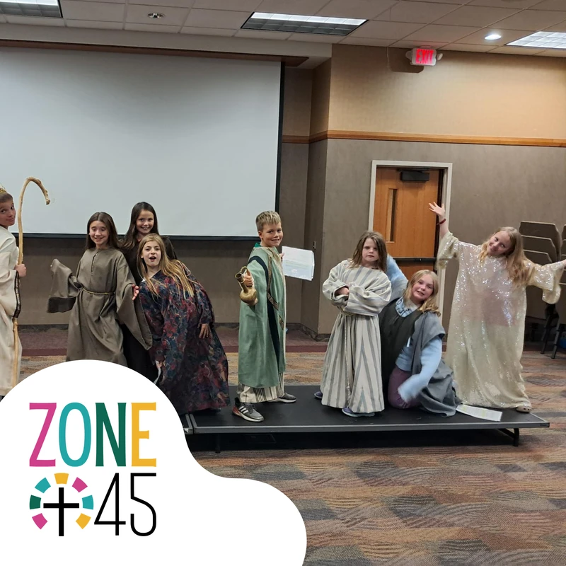 A group of smiling children in biblical costumes pose on a small stage for a play, with a 'ZONE 45' logo in the foreground.