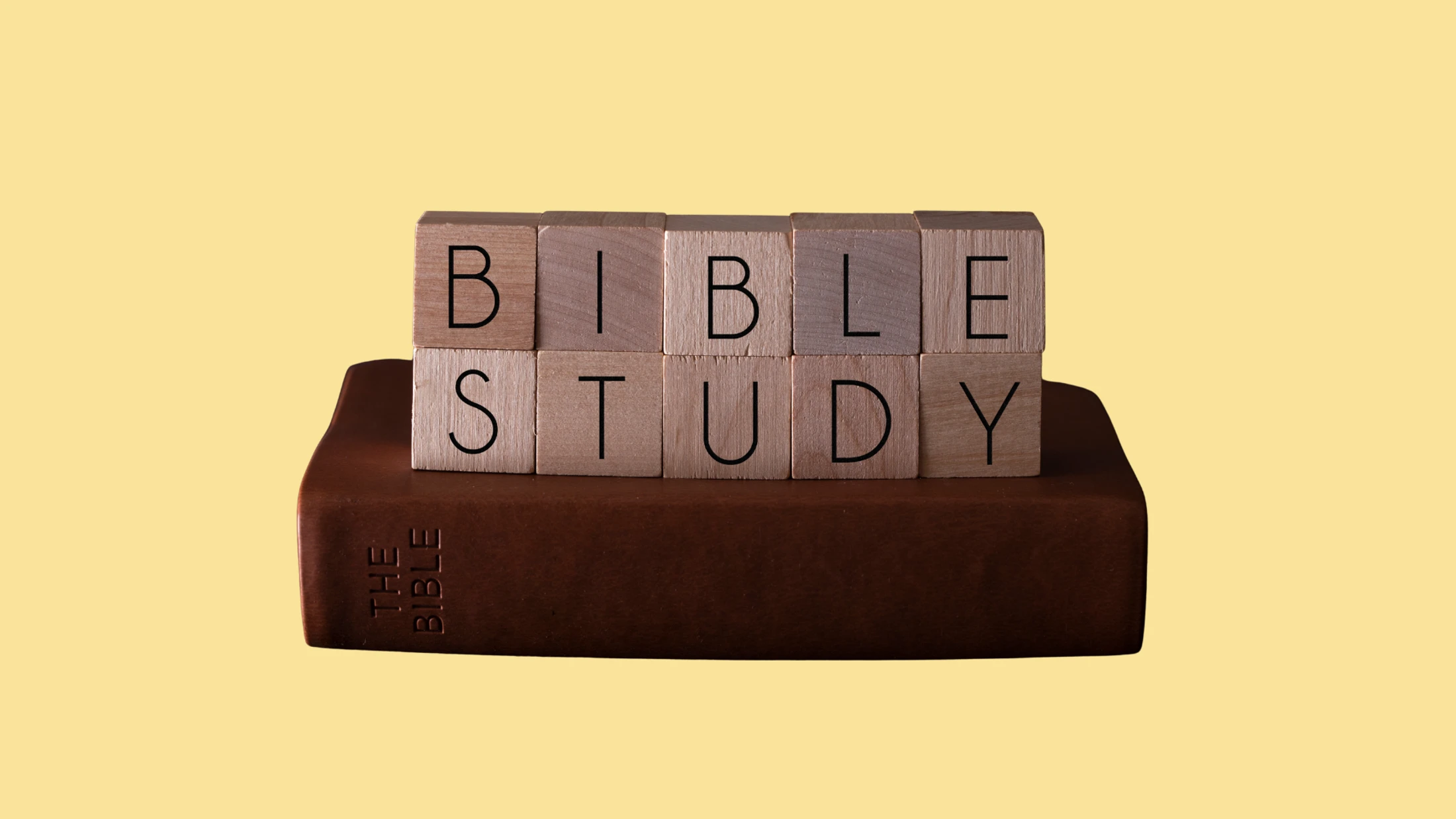 Wooden blocks spelling out 'Bible Study' sit on top of a brown leather Bible against a solid yellow background.