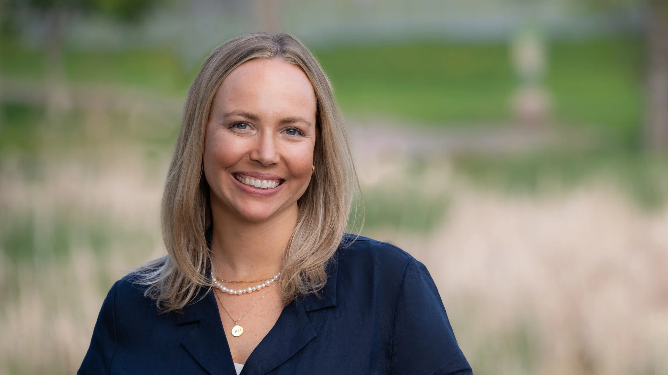 A head and shoulders portrait of a smiling blonde woman wearing a navy blue top and necklaces, standing outdoors with a blurred green and brown background.
