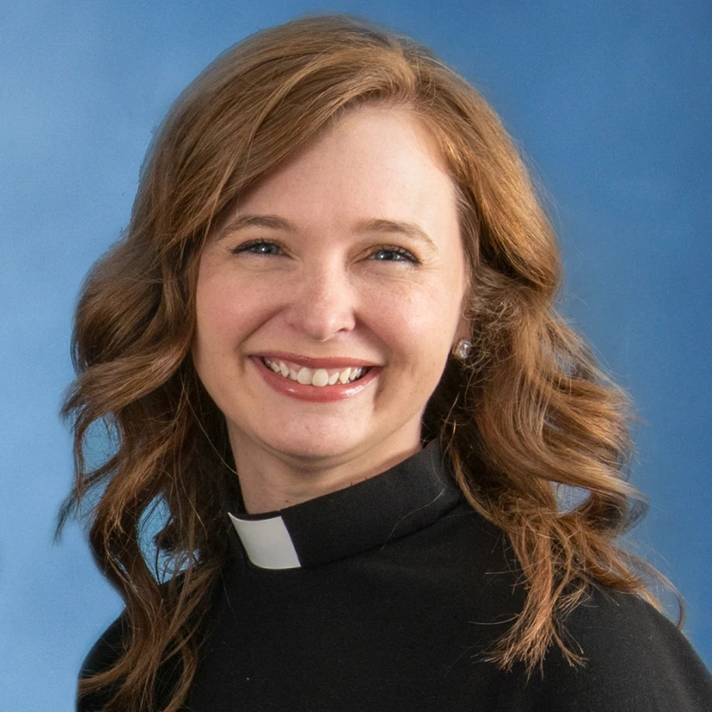 A head and shoulders portrait of a smiling woman with wavy, reddish-brown hair, wearing a black top and a white clerical collar against a blue background.