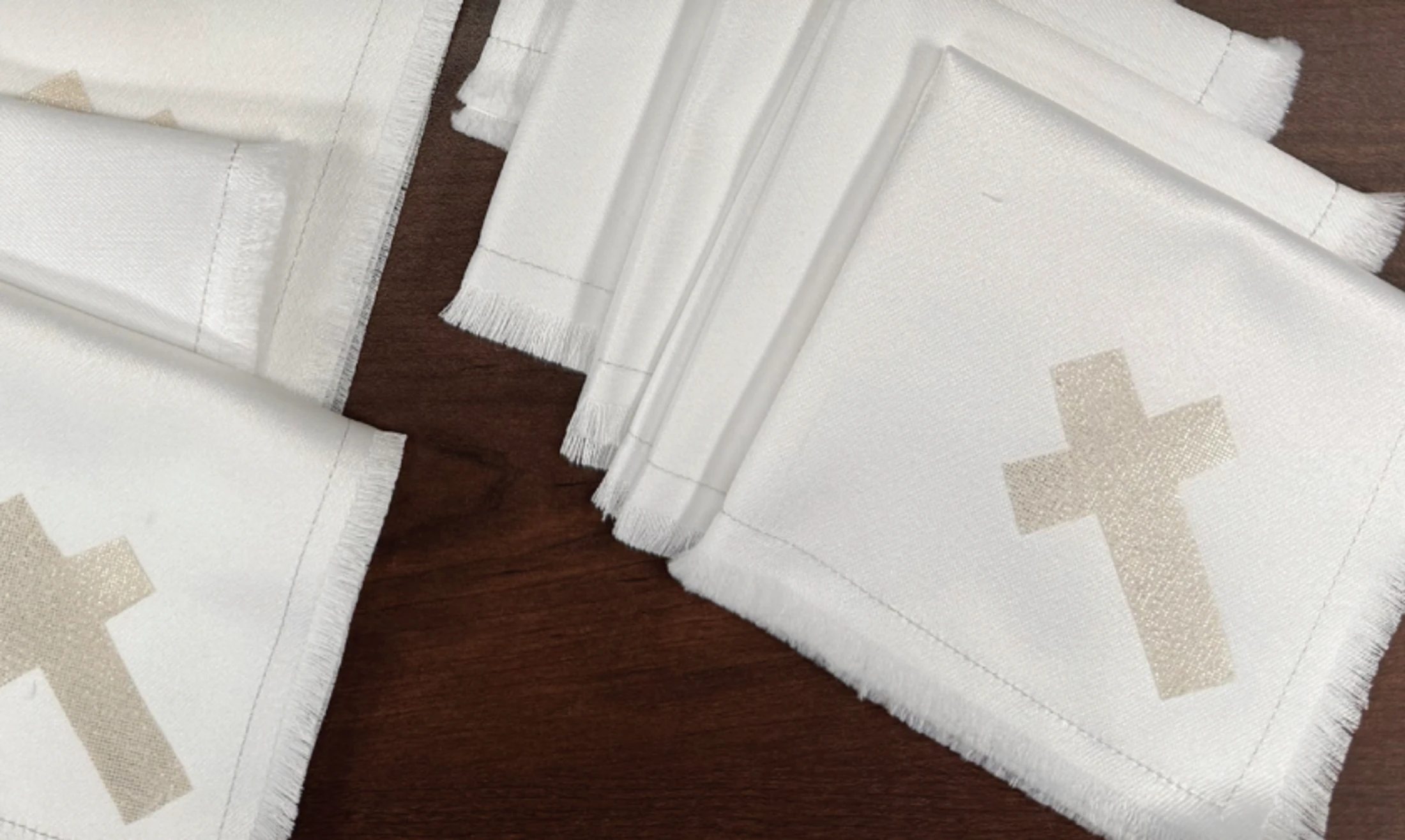 A collection of folded white fabric napkins with frayed edges, each printed with a light gold cross, are arranged on a dark wood surface.