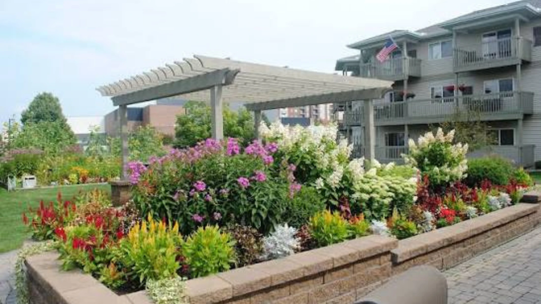 A beautifully landscaped garden with a pergola and a variety of colorful flowers in a raised bed, situated in front of a multi-story apartment building.