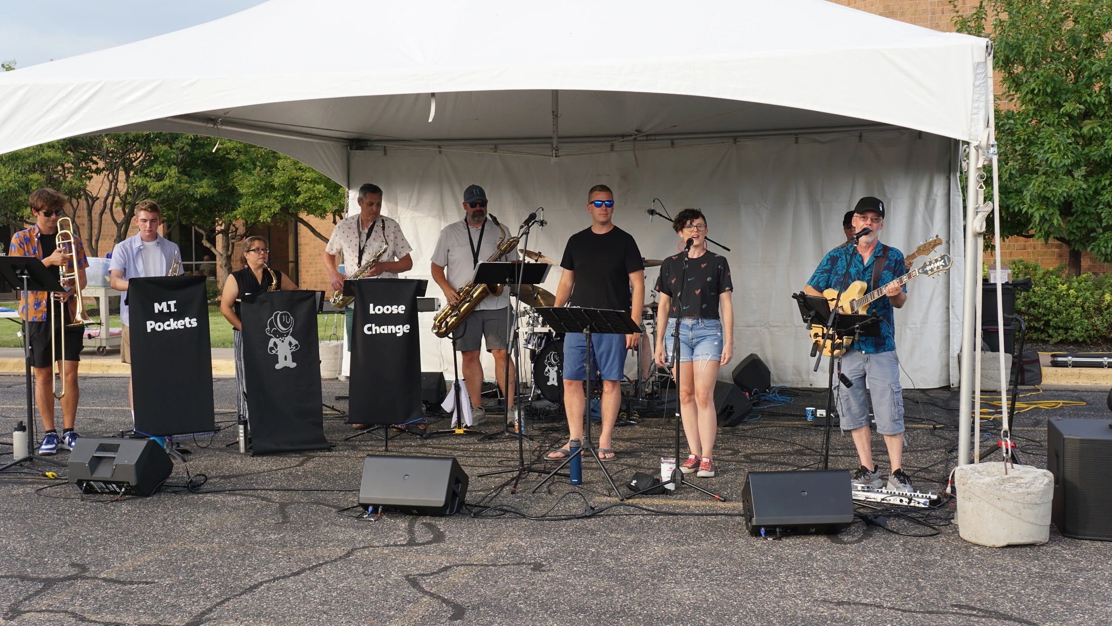 A band with several members, both male and female, performing on a stage under a white tent. They are playing guitars, saxophones, and a trombone.