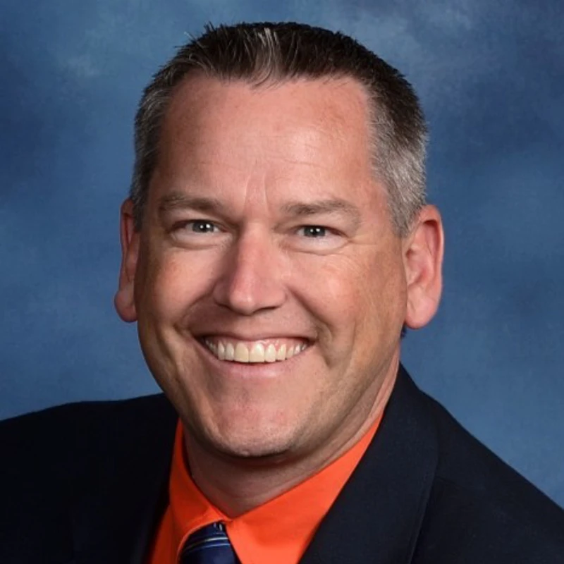 A headshot of a smiling, middle-aged man with short, graying hair. He is wearing a dark suit jacket over a bright orange shirt and a blue tie.