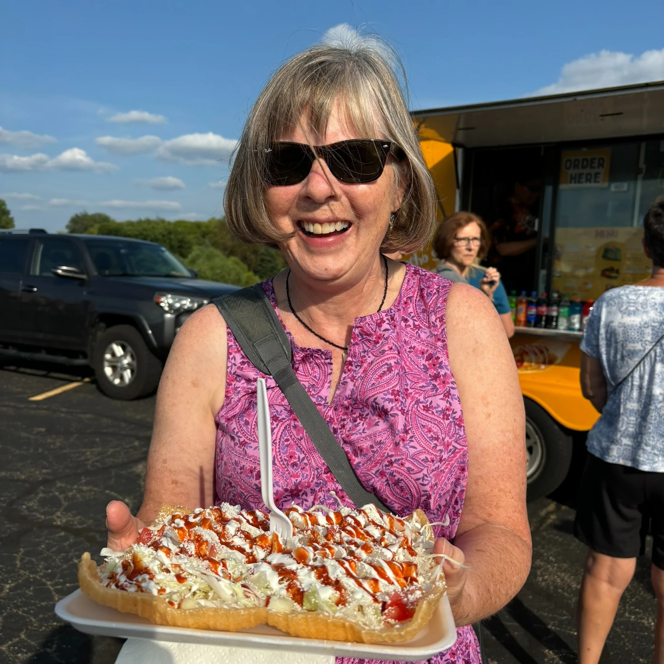 A smiling woman wearing sunglasses holds a large plate of food covered in sauce in front of a yellow food truck on a sunny day.